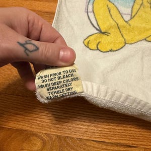 May include: White towel featuring a cartoon character in yellow, blue, and purple. A tag with washing instructions is visible. The tag text reads: WASH PRIOR TO USE, DO NOT BLEACH, WASH DEEP COLORS SEPARATELY, TUMBLE DRY, WARM SETTING.