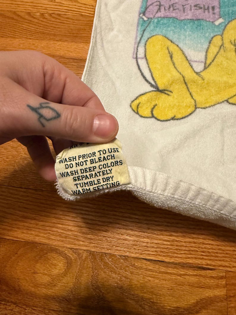 May include: White towel featuring a cartoon character in yellow, blue, and purple. A tag with washing instructions is visible. The tag text reads: WASH PRIOR TO USE, DO NOT BLEACH, WASH DEEP COLORS SEPARATELY, TUMBLE DRY, WARM SETTING.