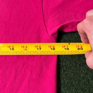 May include: A close-up of a bright pink t-shirt with a yellow measuring tape placed on top of it. The tape measures 15 to 20 inches.