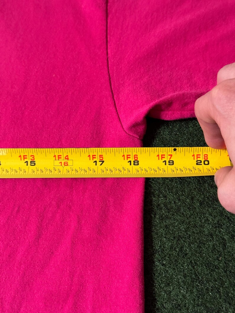 May include: A close-up of a bright pink t-shirt with a yellow measuring tape placed on top of it. The tape measures 15 to 20 inches.