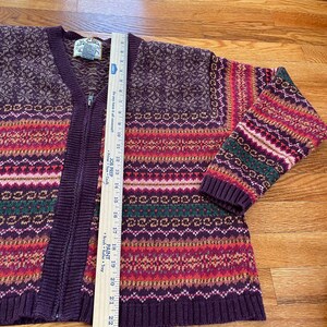 May include: A purple, red, green, and white striped cardigan sweater with a zipper closure. The sweater has a fair isle pattern and is made of a soft, knitted fabric.