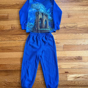 Può includere: Pigiama blu per bambini composto da una maglia a maniche lunghe e pantaloni abbinati. La maglia presenta una grafica di Godzilla su un ponte. Il set è realizzato in tessuto a coste.