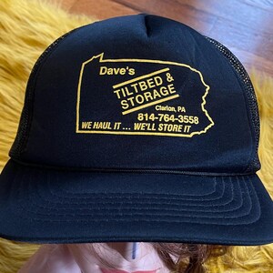 May include: Black trucker hat with a yellow logo that says "Dave's Tiltbed & Storage" with the phone number 814-764-3558 and the address "Clarion, PA". The logo also includes the text "We haul it... We'll store it".