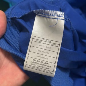 以下が含まれることがあります： 青い衣類のタグのクローズアップ。白いタグには、"100% POLYESTER"や"MADE IN SRI LANKA"などの黒い文字が書かれています。衣類は鮮やかな青色で、端にはリブ編みのテクスチャが見えます。