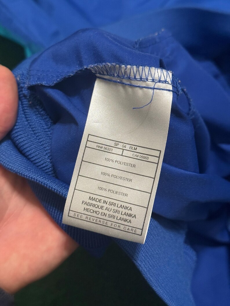 以下が含まれることがあります： 青い衣類のタグのクローズアップ。白いタグには、"100% POLYESTER"や"MADE IN SRI LANKA"などの黒い文字が書かれています。衣類は鮮やかな青色で、端にはリブ編みのテクスチャが見えます。