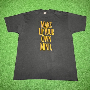 Puede incluir: Camiseta negra con el texto "MAKE UP YOUR OWN MIND" en fuente dorada vertical. La camiseta está colocada sobre una superficie verde.