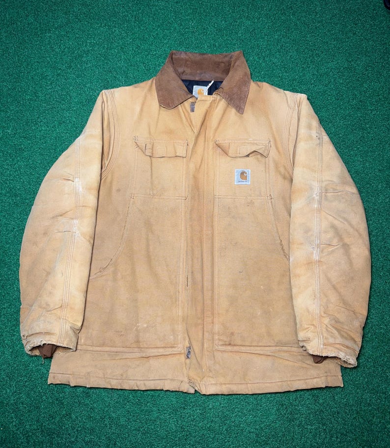 Może przedstawiać: Beżowa kurtka robocza z brązowym kołnierzem sztruksowym i naszywką z logo Carhartt na przedniej kieszeni. Kurtka posiada dwie kieszenie na piersi i dwie dolne kieszenie. Kurtka leży płasko na zielonej powierzchni.