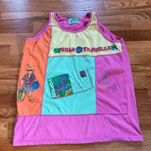 May include: A pink, yellow, orange, and green tank top with a patchwork design. The tank top features a cartoon illustration of a person with a suitcase and the text "WORLD TRAVELLER".