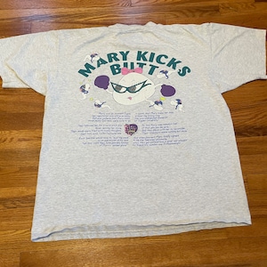 Könnte beinhalten: Graues T-Shirt mit einer Cartoon-Illustration einer Figur namens Mary Kicks Butt. Die Illustration ist in Lila, Rosa und Blau gehalten. Der Text "Mary Kicks Butt" ist in einer lila und blauen Schriftart. Die Rückseite des Shirts enthält eine Beschreibung von Mary Kicks Butt.
