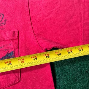 May include: A red t-shirt with a pocket on the left side. The shirt has a black graphic on the pocket and the text "ND" on the front. A measuring tape is partially visible on the right side of the shirt.