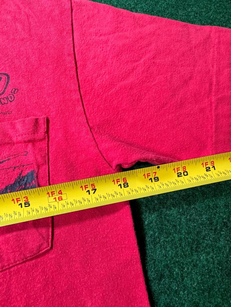 May include: A red t-shirt with a pocket on the left side. The shirt has a black graphic on the pocket and the text "ND" on the front. A measuring tape is partially visible on the right side of the shirt.