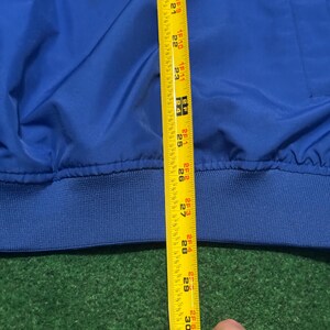 以下が含まれることがあります： 青いジャケットと黄色のメジャーのクローズアップ。テープは約76cmの測定値を示しています。ジャケットはリブ編みの裾と滑らかで光沢のある質感です。