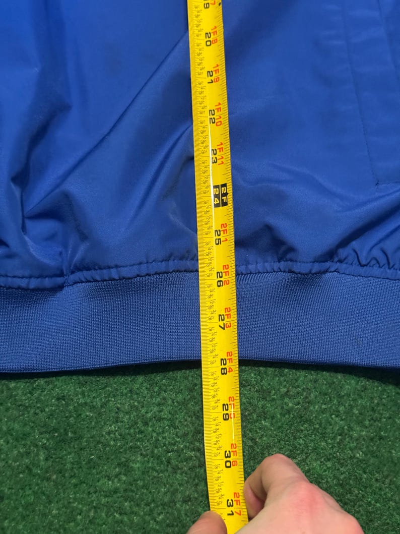 以下が含まれることがあります： 青いジャケットと黄色のメジャーのクローズアップ。テープは約76cmの測定値を示しています。ジャケットはリブ編みの裾と滑らかで光沢のある質感です。