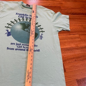 May include: Pale green t-shirt featuring the text "Franklin N Girl Scouts D" and a globe graphic encircled by stick figures. A wooden ruler is placed on the shirt, measuring approximately 74 cm in length.