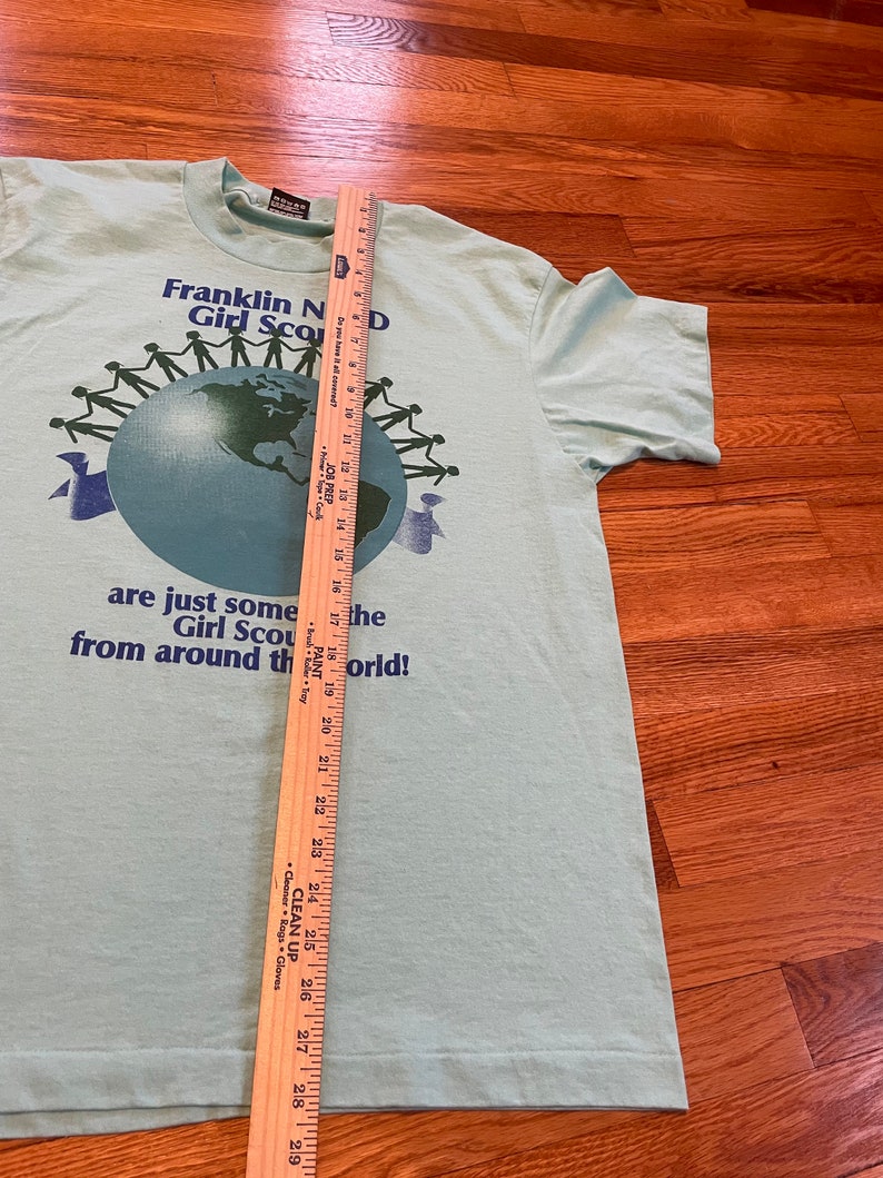 May include: Pale green t-shirt featuring the text "Franklin N Girl Scouts D" and a globe graphic encircled by stick figures. A wooden ruler is placed on the shirt, measuring approximately 74 cm in length.