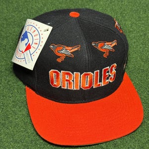 May include: Black and orange baseball cap with the word "Orioles" embroidered on the front. The cap has two small bird logos on the side. The cap is a fitted style.