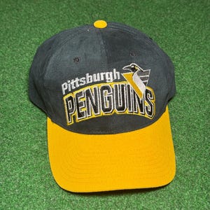 May include: A black and yellow baseball cap with the text "Pittsburgh Penguins" embroidered on the front. The cap features a yellow bill and a yellow button on top. The team logo is also visible.