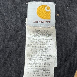 Puede incluir: Primer plano de una etiqueta Carhartt, con el logotipo de la marca en amarillo sobre un fondo blanco. La etiqueta proporciona detalles sobre los materiales de la prenda, incluyendo algod&oacute;n, poli&eacute;ster y nailon. Tambi&eacute;n incluye la talla, el color y las instrucciones de cuidado.
