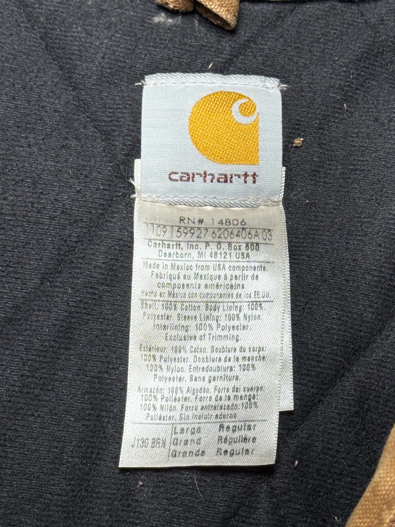Puede incluir: Primer plano de una etiqueta Carhartt, con el logotipo de la marca en amarillo sobre un fondo blanco. La etiqueta proporciona detalles sobre los materiales de la prenda, incluyendo algod&oacute;n, poli&eacute;ster y nailon. Tambi&eacute;n incluye la talla, el color y las instrucciones de cuidado.