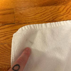 May include: Close-up of a white towel with a dark stain. The towel is folded and has a textured surface. A finger with a black circle tattoo is pointing at the stain.