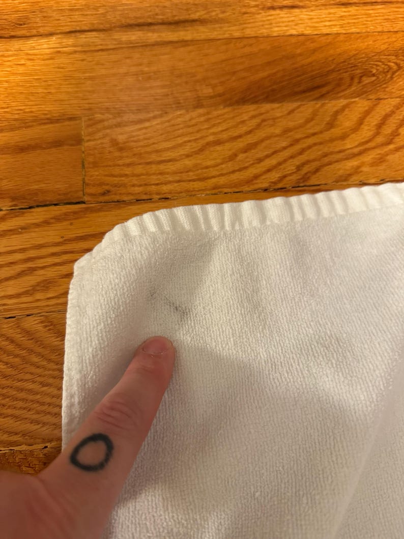 May include: Close-up of a white towel with a dark stain. The towel is folded and has a textured surface. A finger with a black circle tattoo is pointing at the stain.