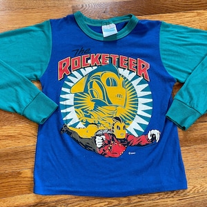 May include: A blue long-sleeve shirt with teal colored sleeves. The shirt features a graphic of The Rocketeer, a superhero character, flying in front of a yellow and red sunburst. The text "The Rocketeer" is printed in red and yellow letters.
