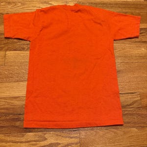 May include: A solid orange t-shirt, viewed from above, with short sleeves and a crew neck. The shirt is made of a soft, textured fabric and is laid flat on a wooden surface. The color is a vibrant, saturated orange.