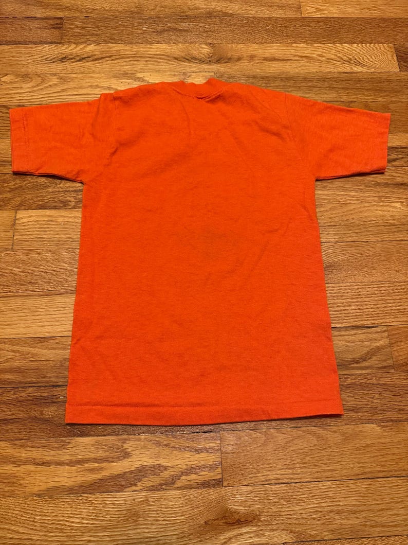 May include: A solid orange t-shirt, viewed from above, with short sleeves and a crew neck. The shirt is made of a soft, textured fabric and is laid flat on a wooden surface. The color is a vibrant, saturated orange.