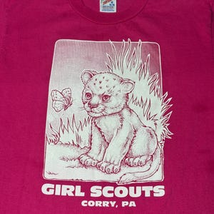 May include: Pink t-shirt with a white graphic of a baby lion cub looking at a butterfly. The graphic is surrounded by grass and the text "GIRL SCOUTS CORRY, PA" is below the graphic.