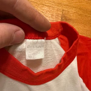 Pu&ograve; includere: Primo piano di una maglietta in stile baseball bianca e rossa. Il colletto e le maniche sono rosse, mentre il corpo &egrave; bianco. Un'etichetta &egrave; visibile all'interno del colletto. La maglietta sembra essere fatta di un materiale morbido, forse vintage.