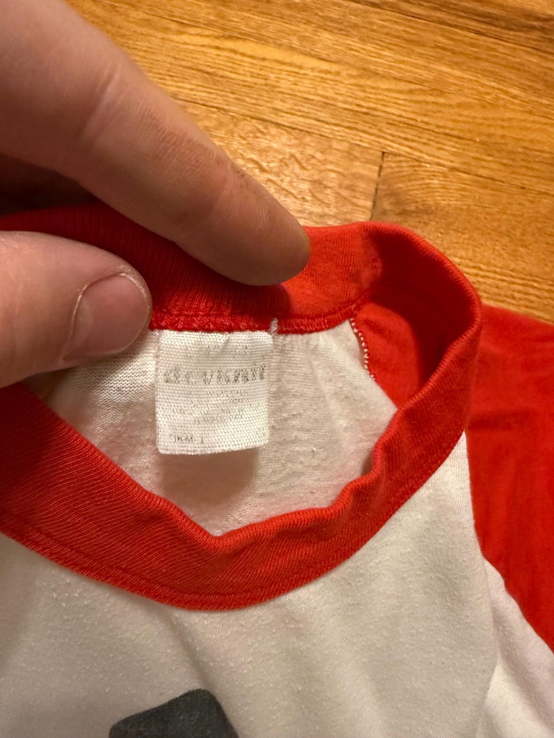 Pu&ograve; includere: Primo piano di una maglietta in stile baseball bianca e rossa. Il colletto e le maniche sono rosse, mentre il corpo &egrave; bianco. Un'etichetta &egrave; visibile all'interno del colletto. La maglietta sembra essere fatta di un materiale morbido, forse vintage.