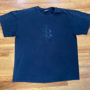 May include: Dark navy blue t-shirt with a faded graphic design on the front. The graphic appears to be a stylized letter "B" with ornate details and text below. The shirt is laid flat on a wooden surface.