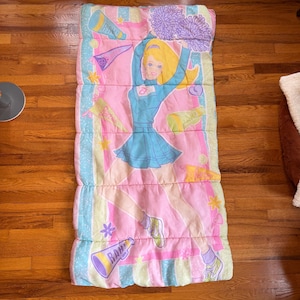 May include: A pink and blue sleeping bag featuring a cartoon cheerleader with blonde hair and a blue uniform. The sleeping bag has a repeating pattern of cheerleading elements, including pom-poms and megaphones. The sleeping bag is rectangular and padded.