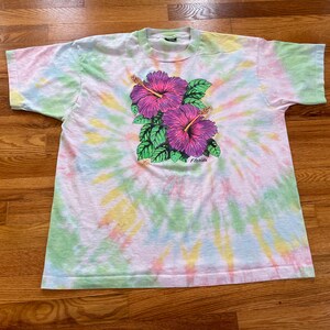 May include: A tie-dye t-shirt with a pink and purple hibiscus flower design. The shirt has a white background with shades of green, yellow, and blue.