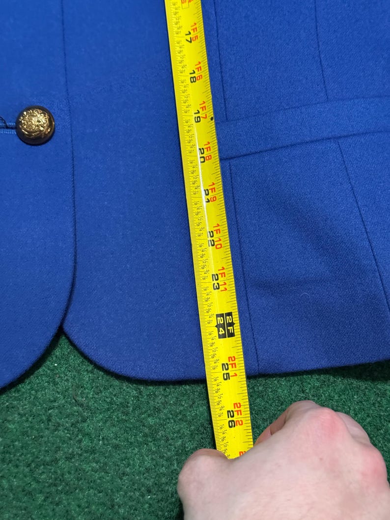 May include: A royal blue blazer with a gold button. The blazer is being measured with a yellow tape measure. The measurement is approximately 66 cm.