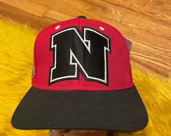 May include: A red and black baseball cap with a large black letter "N" on the front. The cap has a black brim and a white outline around the letter.