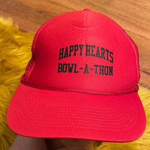 Può includere: Un cappellino da camionista rosso con la scritta "HAPPY HEARTS BOWL-A-THON" in nero. Il cappello ha un retro in rete e una visiera curva. Il cappello è di colore rosso acceso.