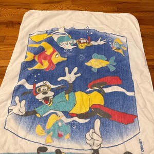 May include: White beach towel featuring Disney characters Goofy, Donald Duck, and Mickey Mouse snorkeling with colourful fish. The towel has a blue background with bubbles and the Disney logo.