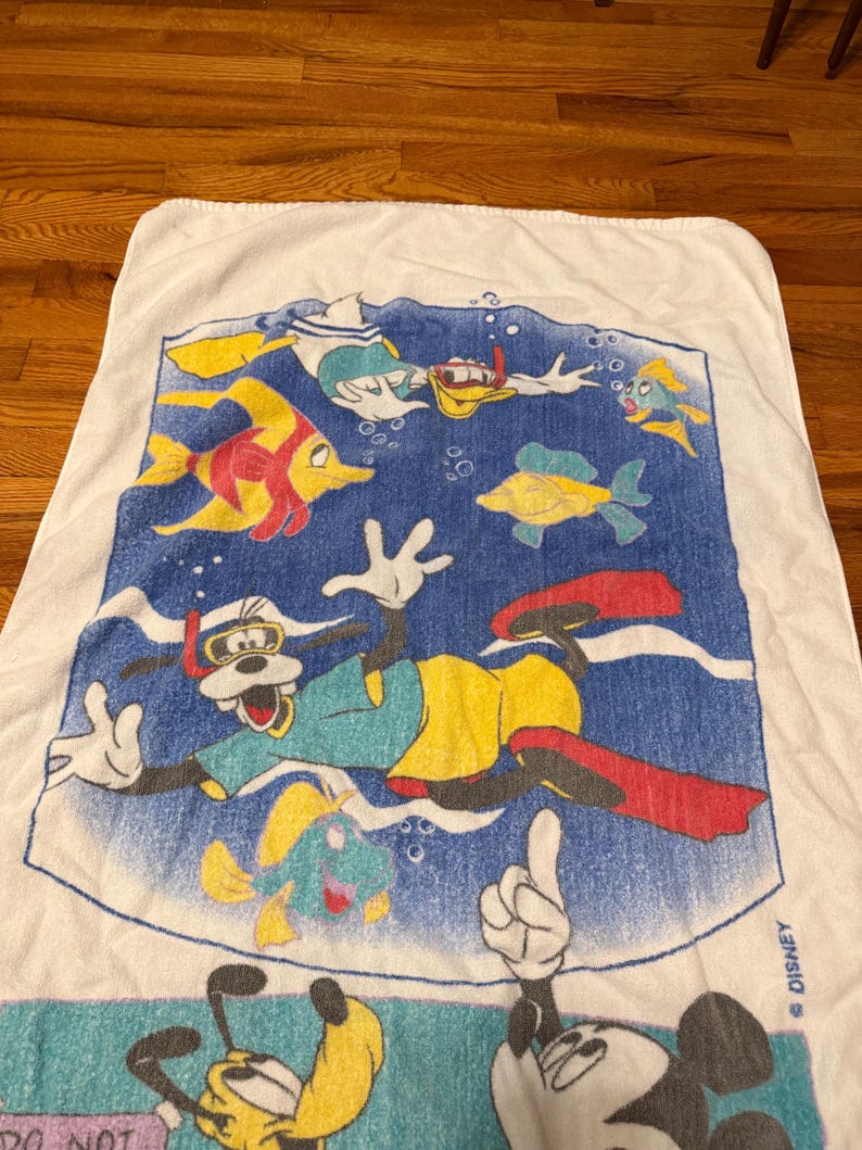May include: White beach towel featuring Disney characters Goofy, Donald Duck, and Mickey Mouse snorkeling with colourful fish. The towel has a blue background with bubbles and the Disney logo.