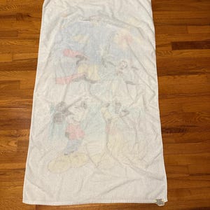 May include: White rectangular beach towel with a faded cartoon graphic of Mickey Mouse, Donald Duck, and Goofy. The towel has a soft, absorbent texture, ideal for use at the beach or pool.