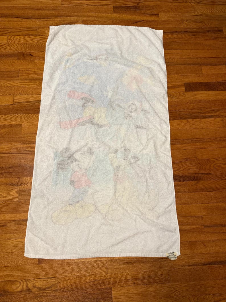 May include: White rectangular beach towel with a faded cartoon graphic of Mickey Mouse, Donald Duck, and Goofy. The towel has a soft, absorbent texture, ideal for use at the beach or pool.