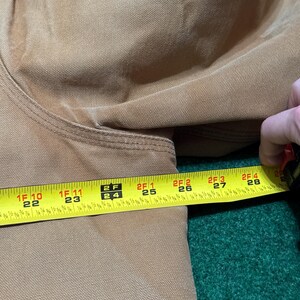 Może przedstawiać: Zbliżenie beżowej tkaniny z miarką pokazującą długość około 56 do 71 cm. Taśma jest ż&oacute;łta z czarnymi oznaczeniami i cyframi. Tkanina wydaje się być trwałym materiałem, prawdopodobnie do odzieży roboczej.