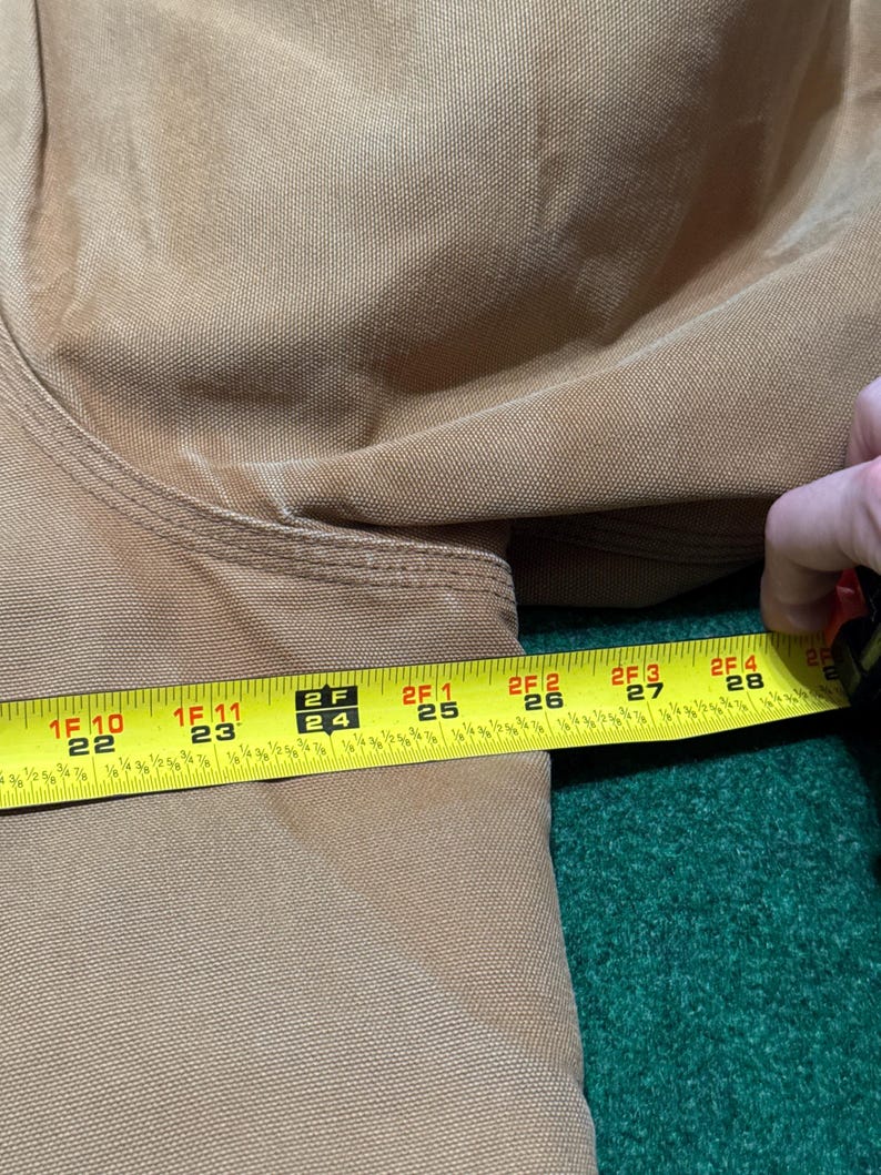 Może przedstawiać: Zbliżenie beżowej tkaniny z miarką pokazującą długość około 56 do 71 cm. Taśma jest ż&oacute;łta z czarnymi oznaczeniami i cyframi. Tkanina wydaje się być trwałym materiałem, prawdopodobnie do odzieży roboczej.