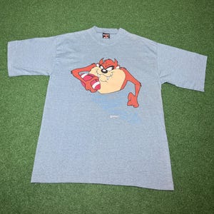 Pode incluir: Camiseta azul claro com um personagem de desenho animado Taz segurando uma bola de futebol americano. O personagem Taz é em tons de vermelho, laranja e bege. A camiseta tem mangas curtas e gola redonda.