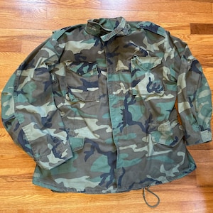 May include: A camouflage jacket with a button-up front and two large pockets on the chest. The jacket has a drawstring at the bottom.