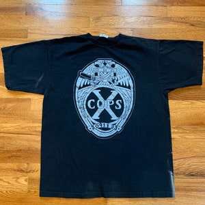 Puede incluir: Camiseta negra con un gráfico blanco de un escudo alado con el texto "X COPS 50911 33" en su interior.