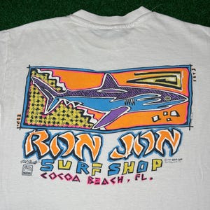 May include: A white t-shirt featuring a graphic print of a shark in blue and purple on an orange background. Below the shark, the text reads RON JON SURF SHOP COCOA BEACH, FL. The design includes yellow and black patterns and colorful lettering.