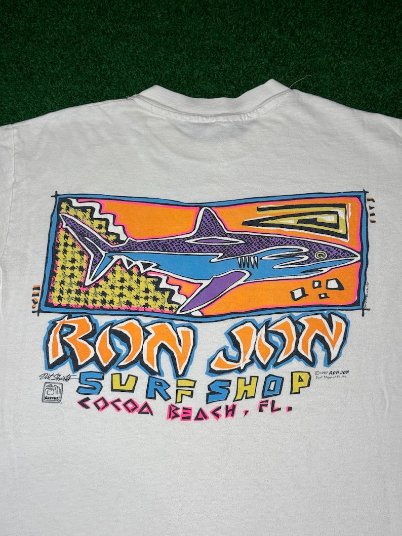 May include: A white t-shirt featuring a graphic print of a shark in blue and purple on an orange background. Below the shark, the text reads RON JON SURF SHOP COCOA BEACH, FL. The design includes yellow and black patterns and colorful lettering.