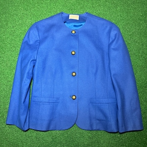 May include: A bright blue blazer with a round neckline and three gold buttons. The jacket has two front pockets and long sleeves. The garment is displayed on a green surface.