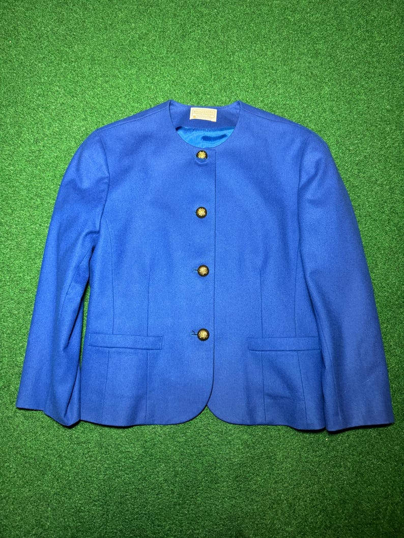 May include: A bright blue blazer with a round neckline and three gold buttons. The jacket has two front pockets and long sleeves. The garment is displayed on a green surface.